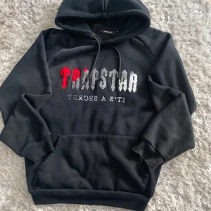 Svart hoodie från Trapstar - Säljer en svart hoodie från Trapstar med broderad logga i rött och grått framtill. Tröjan har huva med dragsko och en stor magficka. Perfekt för dig som gillar streetwear och vill ha något bekvämt och snyggt.