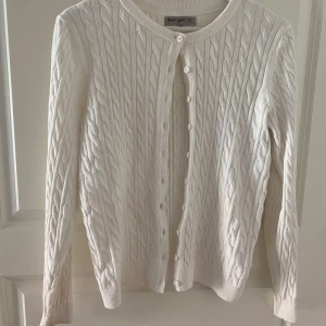 Vit kabelstickad kofta från Basic You - Säljer en stilren vit kabelstickad kofta från Basic You i ekologisk bomull. Koftan har långa ärmar och knappar framtill, perfekt för en klassisk look. Passar till många olika outfits och tillfällen. Köpt från Ullared. Använd 2 gånger.