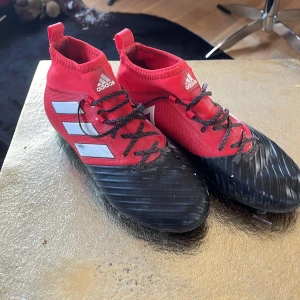 Röd och svart fotbollssko från Adidas - Säljer ett par röda och svarta fotbollsskor från Adidas med vita detaljer och snörning. Skorna har dobbar för spel på gräs. Perfekt för dig som vill sticka ut på planen. Strl 40