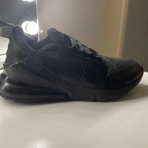 Svarta Nike Air Max 270 sneakers - Säljer ett par helsvarta Nike Air Max 270 sneakers med luftig mesh och synlig Air-dämpning i hälen. Skorna har snörning och en sportig design med diskret Nike-logga på sidan. Perfekta för dig som gillar stilrena och bekväma sneakers.