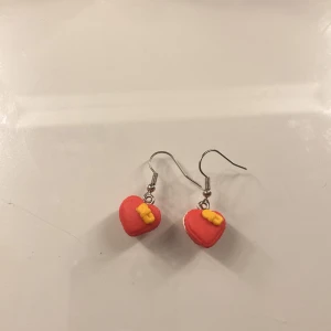 Röda hjärtformade örhängen med rosett - Söta handgjorda örhängen i form av kakor som ser ut som hjärtan med små gula rosetter. Perfekta för att ge en lekfull och färgglad touch till din stil. Hängande modell med metallkrok. Medföljer två stycken ploppar, jag rengör alltid örhängena innan jag skickar!! 