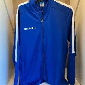 Träningsjacka från Craft - Säljer en blå träningsjacka från Craft med vita detaljer längs axlar och ärmar. Jackan har dragkedja framtill och är långärmad. Perfekt för sport och träning. Märke tryckt på bröstet och bak på ryggen. Helt ny. Ny pris 449