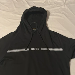 Svart hoodie från BOSS - Säljer en svart hoodie från BOSS med vit logga och två gråa ränder över bröstet. Tröjan har huva med snörning och är perfekt för en avslappnad stil.