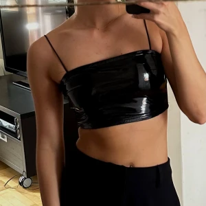 Svart lackinspirerad topp  - Säljer en svart lackinspirerad tubtopp med smala axelband. Kan även användas utan band, se bild! Toppen är croppad och har en tight passform. Materialet är syntet och ger en cool, modern look. Passar till perfekt fest! 