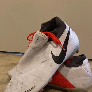 Vita Nike fotbollsskkor - Säljer ett par vita Nike fotbollsskor med svart swoosh och röda detaljer. Skorna har snörning och en strumpliknande öppning för extra stöd. Perfekta för dig som vill sticka ut på planen.