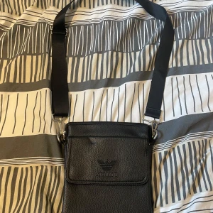 Svart axelväska från Emporio Armani - Snygg svart axelväska från Emporio Armani i skinnimitation med justerbart axelband. Väskan har flera fack med dragkedja och logotyp framtill. Perfekt för att bära det viktigaste med stil.