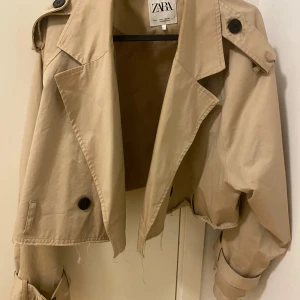 Beige croppad trenchcoat från Zara - Säljer en beige croppad trenchcoat från Zara med stora svarta knappar och bältdetaljer vid ärmsluten. Jackan har klassisk krage och oversized passform, perfekt för en trendig look. Bältet finns även med.