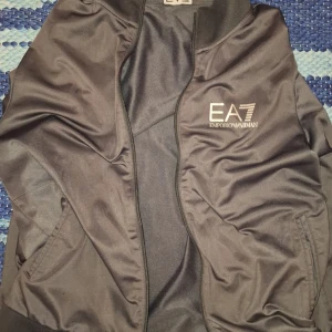 Svart jacka från Emporio Armani EA7 - Snygg svart jacka från Emporio Armani EA7 med logga på bröstet. Jackan har dragkedja framtill, ribbade muddar och två fickor. Perfekt för dig som vill ha en stilren och sportig look.