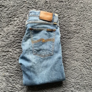 Nudie jeans  - Säljer ett par ljusblå skinny jeans från Nudie Jeans med klassiska orange sömmar och femficksdesign. Jeansen har normal passning i midjan och är tillverkade i denim med dragkedja. Perfekta för dig som gillar en smal siluett. Längd 107cm