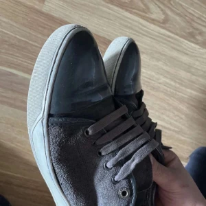 Svarta sneakers från Lanvin med mockadetaljer - Säljer ett par svarta sneakers från Lanvin med ovandel i mocka och tåparti i blankt material. Skorna har snörning och vit sula. Klassisk och stilren design som passar till mycket.