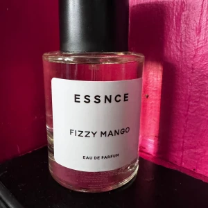ESSNCE Fizzy Mango Eau de Parfum - En fräsch och fruktig parfym från ESSNCE med doft av mango. Flaskan är rund och genomskinlig med svart lock och vit etikett. Perfekt för dig som gillar tropiska och söta dofter.