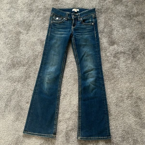 Blå bootcut jeans med fickdetaljer - Säljer ett par klassiska blå jeans med bootcut passform. Jeansen har fram- och bakfickor med snygga knappdetaljer och kontrastsömmar. Perfekta för dig som gillar en tidlös stil med lite utsvängda ben.