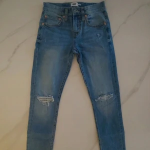 Blå slitna jeans med hål på knäna - Säljer ett par blå jeans med slitningar och hål på båda knäna. Klassisk femficksmodell med knappgylf och orangea sömmar. Jeansen har en smal passform och är perfekta för en avslappnad stil.