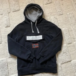 Svart hoodie från Napapijri - Säljer en svart hoodie från Napapijri med grå insida på huvan och justerbara snören. På bröstet finns en stor ficka med Napapijri-logga och en norsk flagga. Perfekt för en avslappnad och snygg stil.