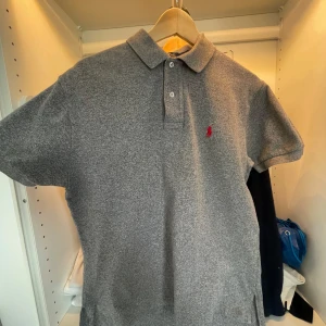 Grå pikétröja från Polo Ralph Lauren - Klassisk grå pikétröja från Polo Ralph Lauren med liten röd broderad logga på bröstet. Tröjan har krage, knappar vid halsen och korta ärmar. Perfekt för en stilren och avslappnad look.