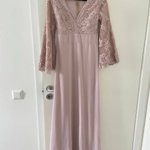 Ljusrosa långklänning med spetsärmar - Elegant ljusrosa långklänning med v-ringning och vida, långärmade spetsärmar. Överdelen har vackra spetsdetaljer och kjolen faller rakt ner i ett mjukt tyg. Dragkedja i ryggen för enkel påklädning.Ljusrosa lång klänning,  Märke:NLY Trend  Bra skickt 