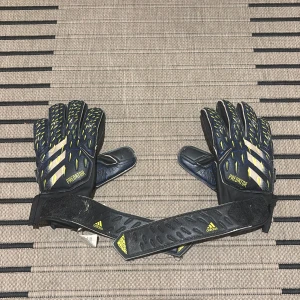 Adidas Predator målvaktshandskar - Säljer ett par svarta Adidas Predator målvaktshandskar med gula och blå detaljer samt silvriga ränder. Handskarna har justerbara kardborreband och är designade för fotbollsmålvakter. Kontakta vid frågor, pris:80kr 