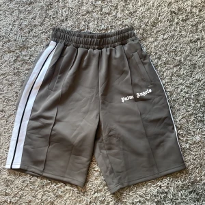 Grå shorts från Palm Angels med vita ränder - Snygga grå shorts från Palm Angels med vita sidoränder och logga tryckt på vänster ben. Shortsen har elastisk midja och sidofickor. Perfekta för en avslappnad och trendig stil.