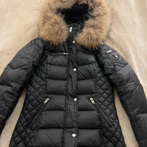 Svart dunjacka med päls från Rockandblue - Säljer en svart lång dunjacka från Rockandblue med snygg fuskpäls runt huvan. Jackan har dragkedjor, knappar och flera fickor samt quiltade detaljer på sidor och ärmar. Perfekt för kalla dagar och riktigt stilren look.