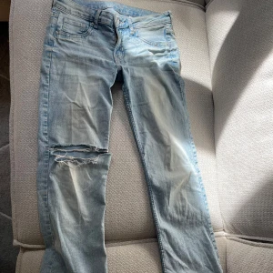 Ljusblå låg midjade slitna jeans - Säljer ett par ljusblå jeans med slitningar och hål på vänster ben. Jeansen har en utsvängd passform och är lågmidjade perfekt för en avslappnad stil. Storlek 40 men skulle snarare säga att dem är storlek 38.