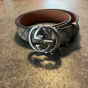 Gucci bälte med silverfärgat spänne - Säljer ett Gucci bälte med klassiskt GG-mönster i grått och svart. Bältet har ett stort, rundat spänne i silver med dubbla G:n. Insidan är ljusbrun och materialet är tåligt med exklusiv känsla. 105 cm