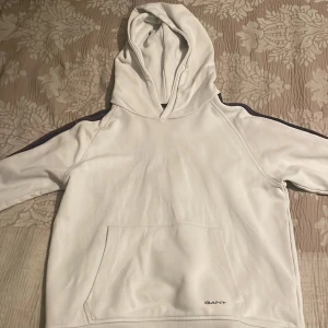 Vit hoodie från GANT med logga på ärmen - Säljer en vit hoodie från GANT med blå logga längs ärmen och broderad GANT-text vid fickan. Tröjan har huva och magficka, samt ribbade muddar. Perfekt för en avslappnad och sportig stil.