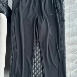 Svarta träningsbyxor från Nike - Svarta träningsbyxor från Nike med dragkedja vid fickorna och diskret logga på låret. Byxorna har normal passform och är tillverkade i syntetmaterial, perfekta för träning eller sportiga dagar.