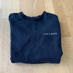 Mörkblå sweatshirt från Lyle & Scott - Mörkblå sweatshirt från Lyle & Scott med rund halsringning och ribbade muddar. Diskret logotyp broderad på bröstet och en gul broderad detalj på ärmen. Perfekt för en avslappnad stil.