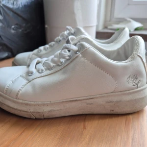 Vita sneakers med silvriga detaljer - Säljer ett par vita sneakers med silvriga detaljer på hälen och plösen. Skorna har en ros på. Men båda skorna är sönder i mitten (bild 5) och bakkapporna är slitna (bild 2). Perfekta för en stilren och enkel look.