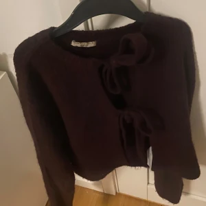 Mörklila stickad kofta med rosetter - Säljer en mörklila stickad kofta från Gina  med två stora rosetter framtill. Koftan har lång ärm och är supermjuk och mysig, perfekt för lager-på-lager. Passar dig som gillar unika detaljer!
