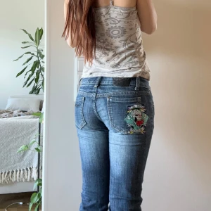 Ed Hardy jeans med broderad dödskalle - Säljer ett par blå jeans från Ed Hardy med en stor broderad dödskalle och färgglada detaljer på bakfickan. Klassisk femficksmodell med normal passform och låg midja. Snygga kontrastsömmar och cool knapp med logga framtill. Strl S