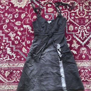 Mörkgrå omlottklänning från Taboo - Säljer en mörkgrå omlottklänning från Taboo i storlek 34. Klänningen har smala axelband som knyts, v-ringning och ett knytband i midjan. Perfekt för sommardagar och enkel att styla med sneakers eller sandaler.