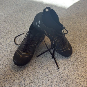 Svarta Nike fotbollsskor med guldiga detaljer - Säljer ett par svarta Nike fotbollsskor med guldiga och vita detaljer samt snörning. Skorna har högre skaft och mönstrad sula för bra grepp på planen. Swoosh-logga på sidorna och diskreta grafiska linjer.