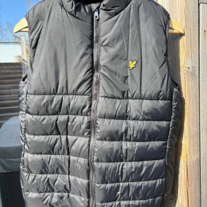 Svart dunväst från Lyle & Scott Junior - Snygg svart dunväst från Lyle & Scott Junior med gul logotyp på bröstet. Västen har quiltad design och dragkedja framtill. Perfekt för lager-på-lager och passar till många olika stilar och nu till våren och sommaren.                                                                  Nypris: ca 750kr                                                                 Pris kan diskuteras vid snabb affär 