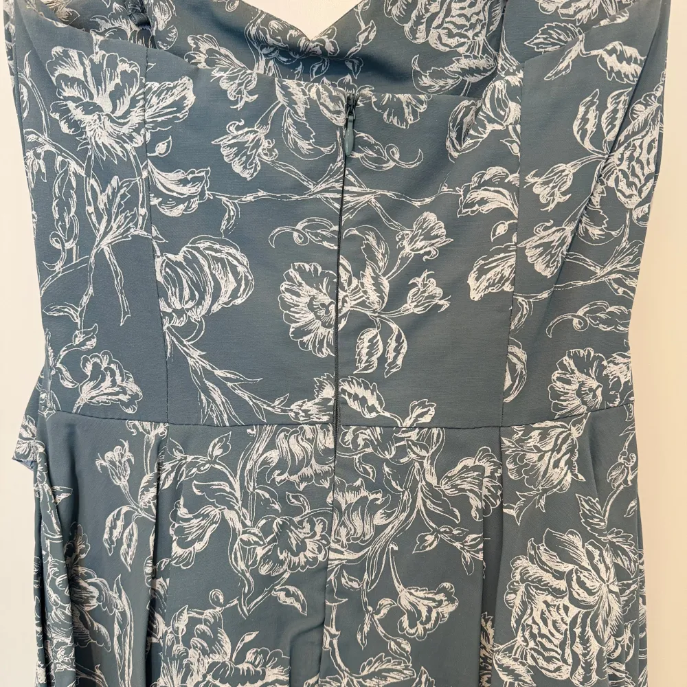 Elegant klänning i en dämpad blå nyans med vackert blommönster i vitt. Klänningen har en v-ringad hals och ett knytband i midjan som ger en smickrande passform. Perfekt för sommarens festligheter.. Mekot.
