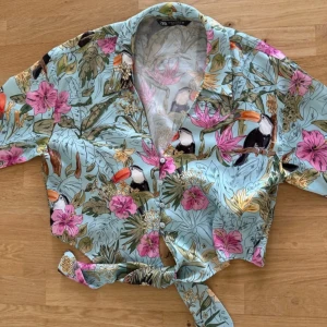 Blommig kortärmad skjorta från Zara - Snygg blommig skjorta från Zara med tropiskt mönster i grönt, rosa och orange. Skjortan har korta ärmar och knytning framtill, perfekt för en somrig look. Tillverkad i ett lätt och luftigt material.