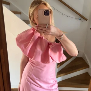 Rosa klänning med volangdetalj - Säljer en söt rosa klänning med en stor volangdetalj över axeln. Klänningen har en tight passform och är perfekt för sommarens fester. Den är axelbandslös och har en elegant design.