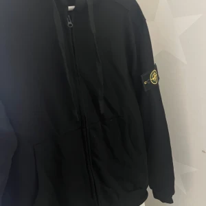 Svart  Stone Island tröja  - Säljer en stilren svart  tröja från Stone Island med dragkedja och deras ikoniska märke på ärmen. Perfekt för en cool och avslappnad look.