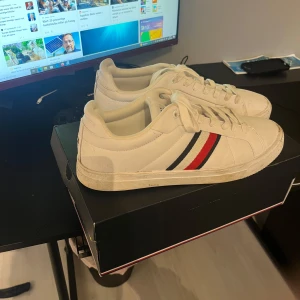 Vita sneakers från Tommy Hilfiger - Säljer ett par vita sneakers från Tommy Hilfiger med klassiska ränder i rött och blått på sidan. Skorna har snörning och en rund tå, vilket ger en stilren look. Perfekta för en casual stil.