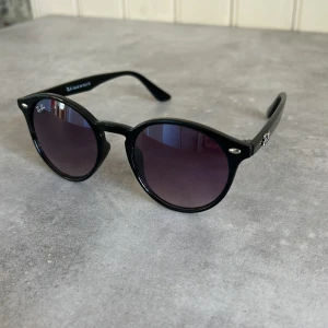 Svarta solglasögon från Ray-Ban - Snygga svarta solglasögon från Ray-Ban med rund form och mörka linser. Perfekta för en stilren look. De har en klassisk design med Ray-Ban-loggan på sidan av bågarna.