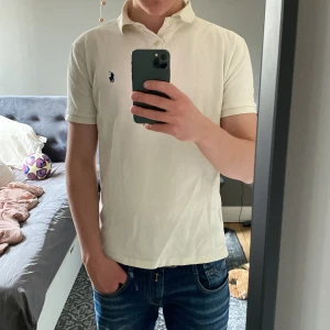 Vit pikétröja från Ralph Lauren - Säljer en klassisk vit pikétröja från Ralph Lauren med den ikoniska loggan broderad på bröstet. Tröjan har korta ärmar och en stilren krage. Perfekt för en avslappnad men stilfull look.