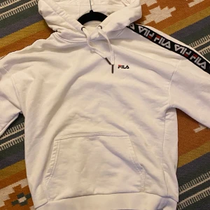 Vit hoodie från FILA - Säljer en snygg vit hoodie från FILA med logotyp på bröstet och längs ärmarna. Den har en klassisk känguruficka och justerbar huva med dragsko. Perfekt för en avslappnad stil.