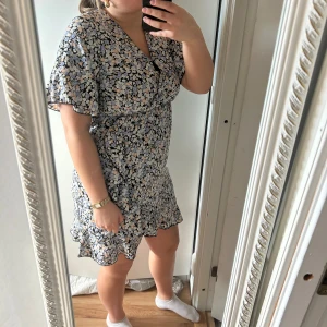 Blommig klänning med volangdetaljer - Charmig blommig klänning med volangdetaljer och v-ringning. Klänningen har korta ärmar och en omlott-design som ger en feminin touch. Perfekt för sommarens alla tillfällen.