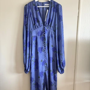 Positano dress Adoore - Säljer en elegant blå klänning från Adoore med vackert blommönster. Klänningen har långa ärmar och en v-ringad design med knappar framtill. Aldrig använd med prislapp kvar.