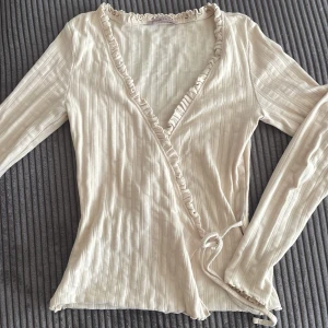 Beige omlottopp med volangdetaljer - Säljer en stilren beige omlottopp med volangdetaljer längs den v-ringade halsen. Toppen har långa ärmar och ett knytband i midjan för en justerbar passform. Perfekt för en chic och avslappnad look.