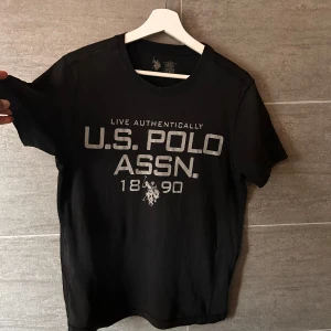Svart t-shirt från U.S. Polo Assn. - Snygg svart t-shirt från U.S. Polo Assn. med tryck framtill. Tillverkad i en mjuk blandning av bomull och polyester för en bekväm passform. Perfekt för en avslappnad stil.