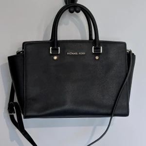 Svart axelväska/handväska från Michael Kors - Fin väska från MK som jag tyvärr inte använder så mycket. Den är köpt på sellpy för en ganska dyr peng och jag vet att de äkthetskontrollerar sina varor så den ska vara äkta (men jag har tyvärr intet orginalkvitto att visa upp förutom mitt från sellpy då. Kan bäras antingen som en handväska eller axelremsväska och axelremmen kan även tas av. Ena fästet till axelremmen är lagat (se bild). Det är lite slitage på piggarna som är på botten. I övrigt inga defekter. Modellen heter AI-1304.