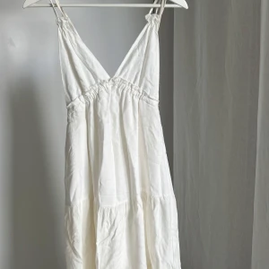 Vit klänning från Bershka - Säljer en vit klänning från Bershka med justerbara axelband och v-ringad design. Klänningen har en luftig passform och är perfekt för varma dagar. Tillverkad i ett lätt och bekvämt material.