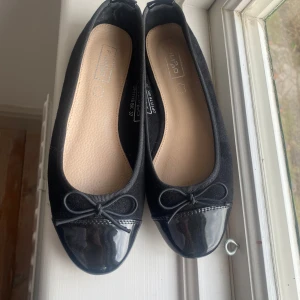 Svarta ballerinaskor med lackdetaljer - Snygga svarta ballerinaskor med lackdetaljer på tån och en söt rosett framtill. Perfekta för en stilren look. Ganska små i storleken skulle säga att storleken är 36/37.