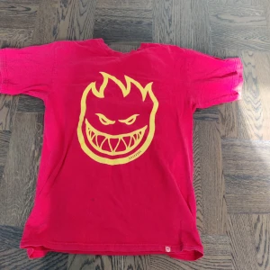Röd Spitfire t shirt - Säljer en röd Spitfire t-shirt med ett gult, eldsflammigt ansikte tryckt på framsidan. T-shirten har korta ärmar och en rund halsringning. Perfekt för en avslappnad stil eller streetwear-look. Väldigt litet håll under ena armen. Passar bra till jeans eller shorts. Skicka ett meddelande vid intresse!